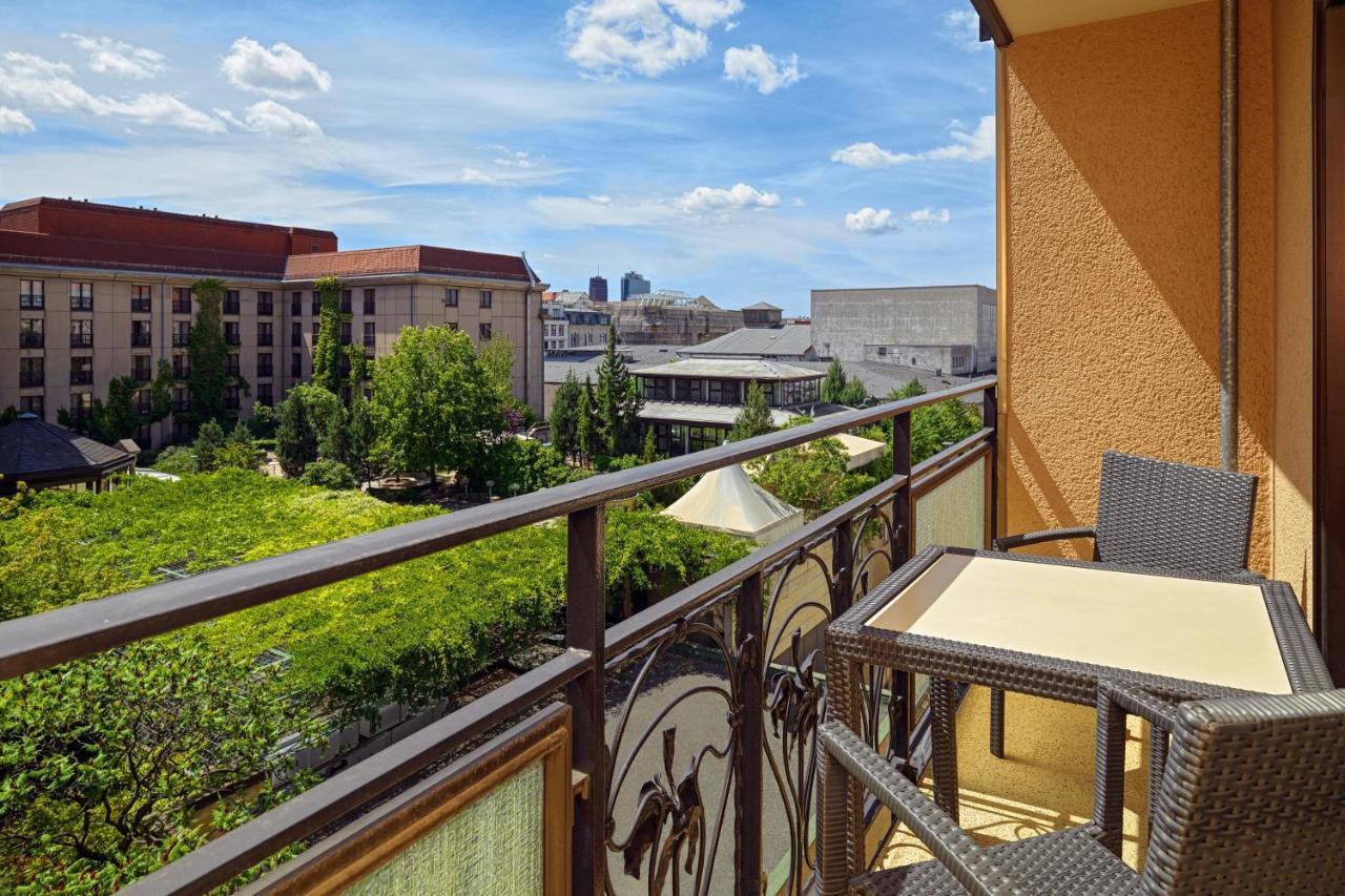 فندق ذا ويستن جراند، برلين المظهر الخارجي الصورة The photo shows a balcony with a view overlooking a vibrant urban scene. There is a railing with decorative design, and a small table with two chairs on the balcony. The area below features greenery and trees, while buildings are visible in the backg